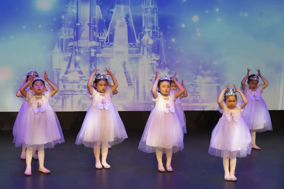 Six young dancers in white dresses performing on stage in front of a castle backdrop