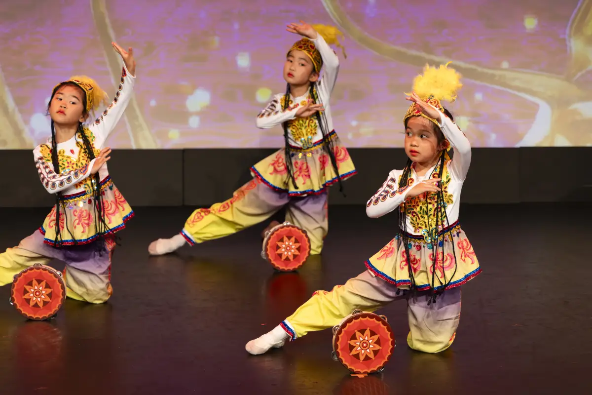 Three young dancers in ornate costumes posing with decorative drums during a stage performance