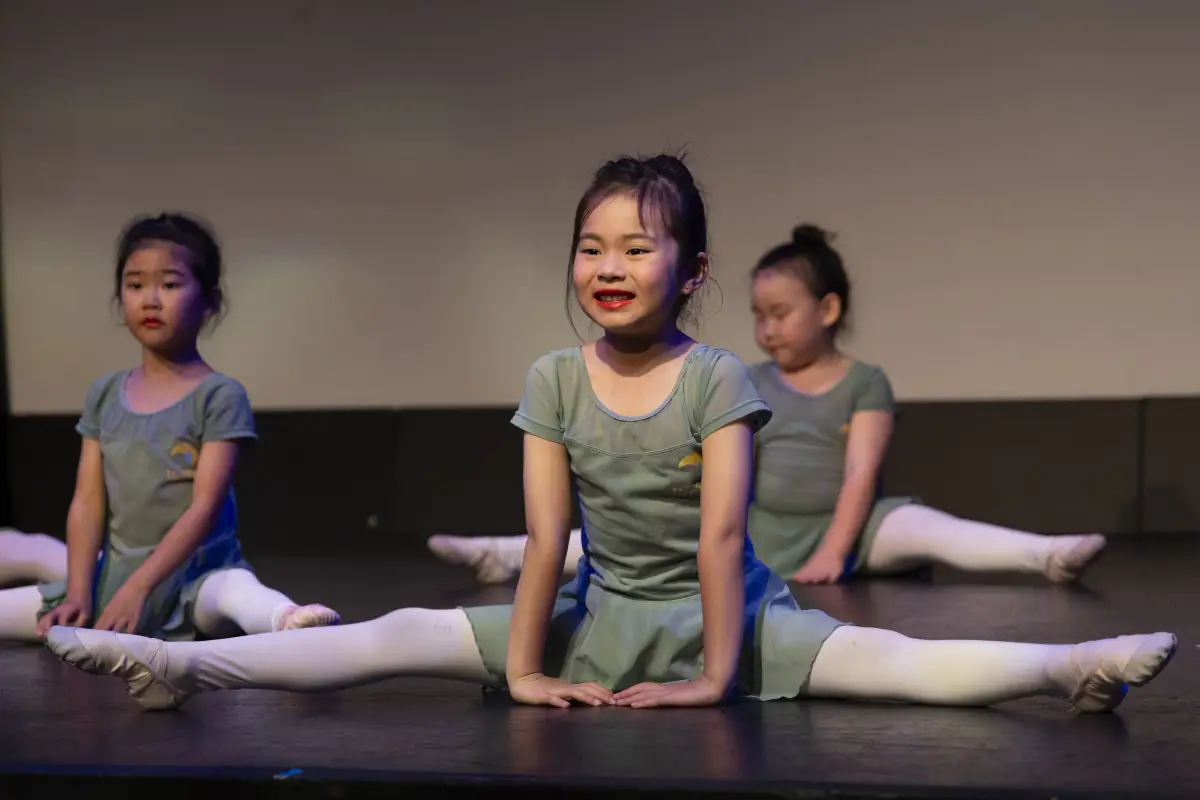 Young dancer in a sage green costume performing the splits on stage with two students behind her