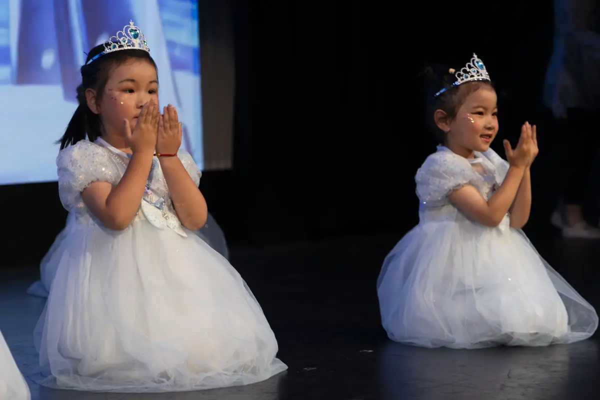 Two young dancers in white tulle dresses and tiaras kneeling on stage during a recital number